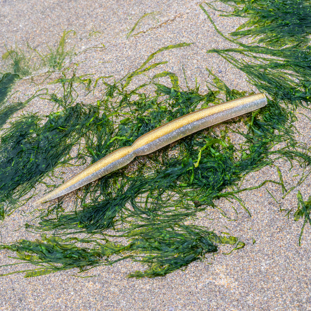 A realistic plastic fishing lure shaped like a small shrimp or bait, resting on green seaweed scattered on sandy beach ground.