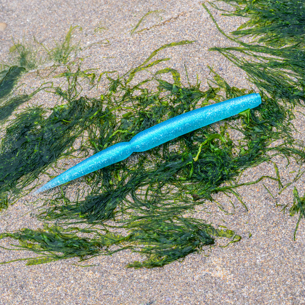 A bright blue glitter soft plastic fishing lure with a segmented body and a curved tail, resting on green seaweed over sandy beach ground.
