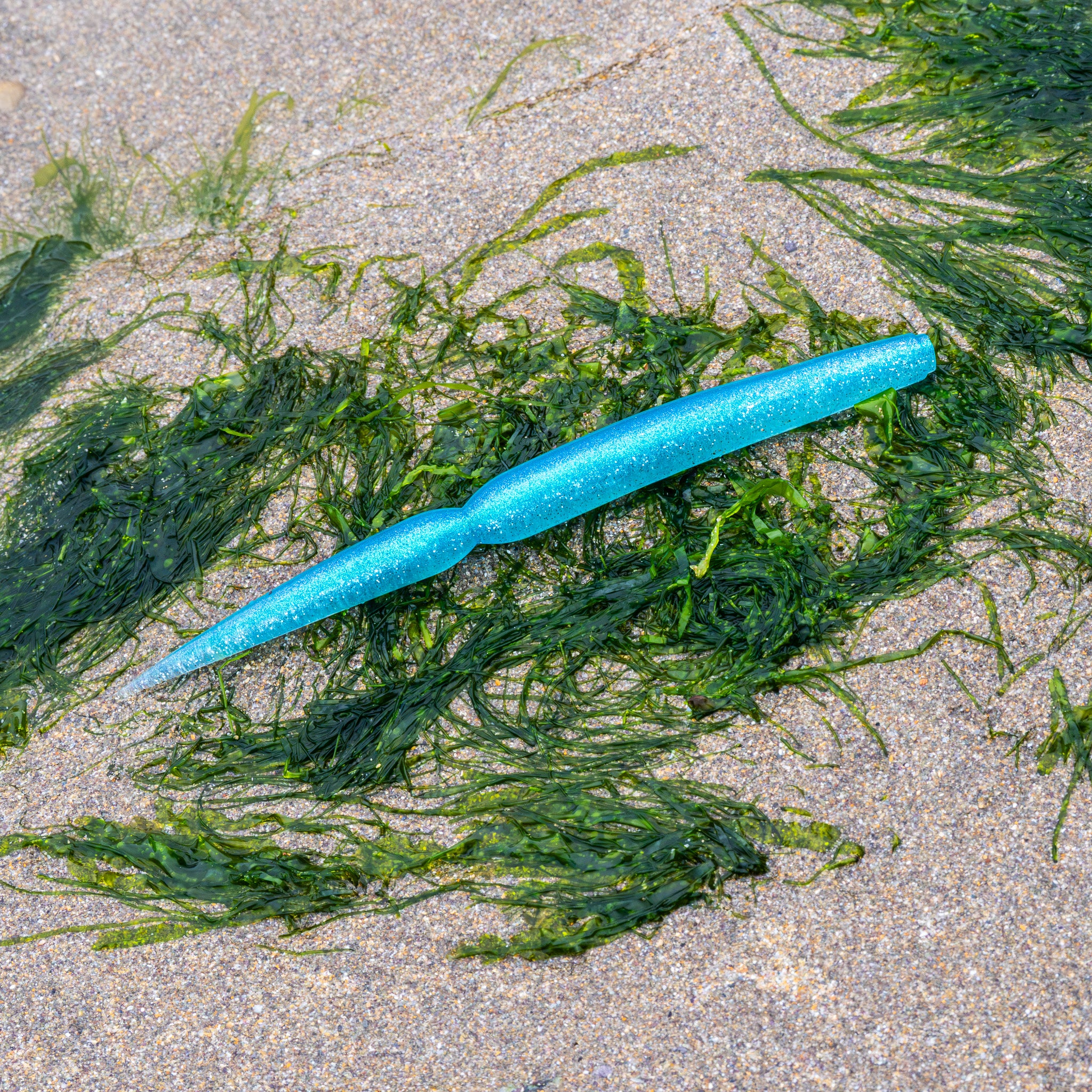 A bright blue glitter soft plastic fishing lure with a segmented body and a curved tail, resting on green seaweed over sandy beach ground.
