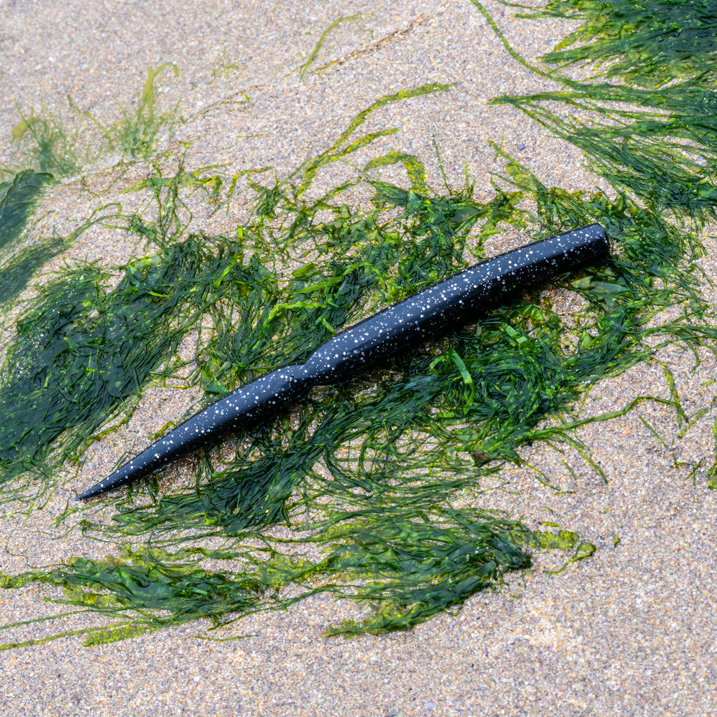 A black soft plastic fishing lure with white glitter flakes, laid diagonally on a sandy beach surrounded by green seaweed.