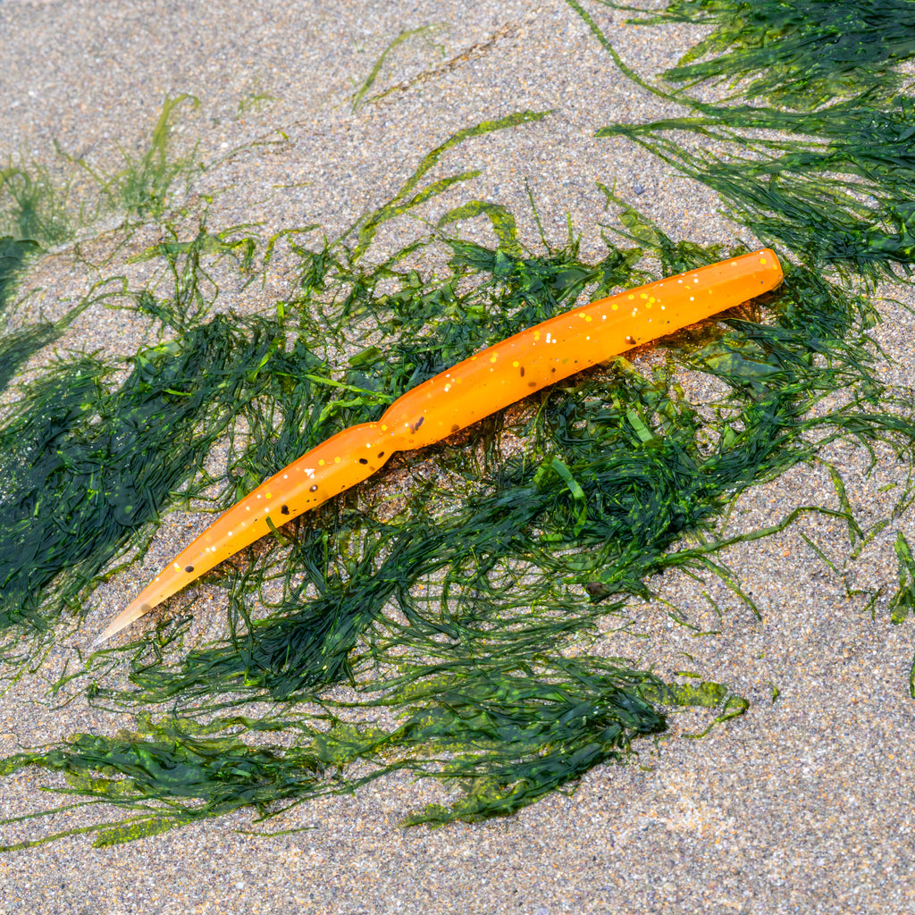 An orange soft plastic fishing lure with black glitter flecks, shown on a sandy beach surrounded by green seaweed.