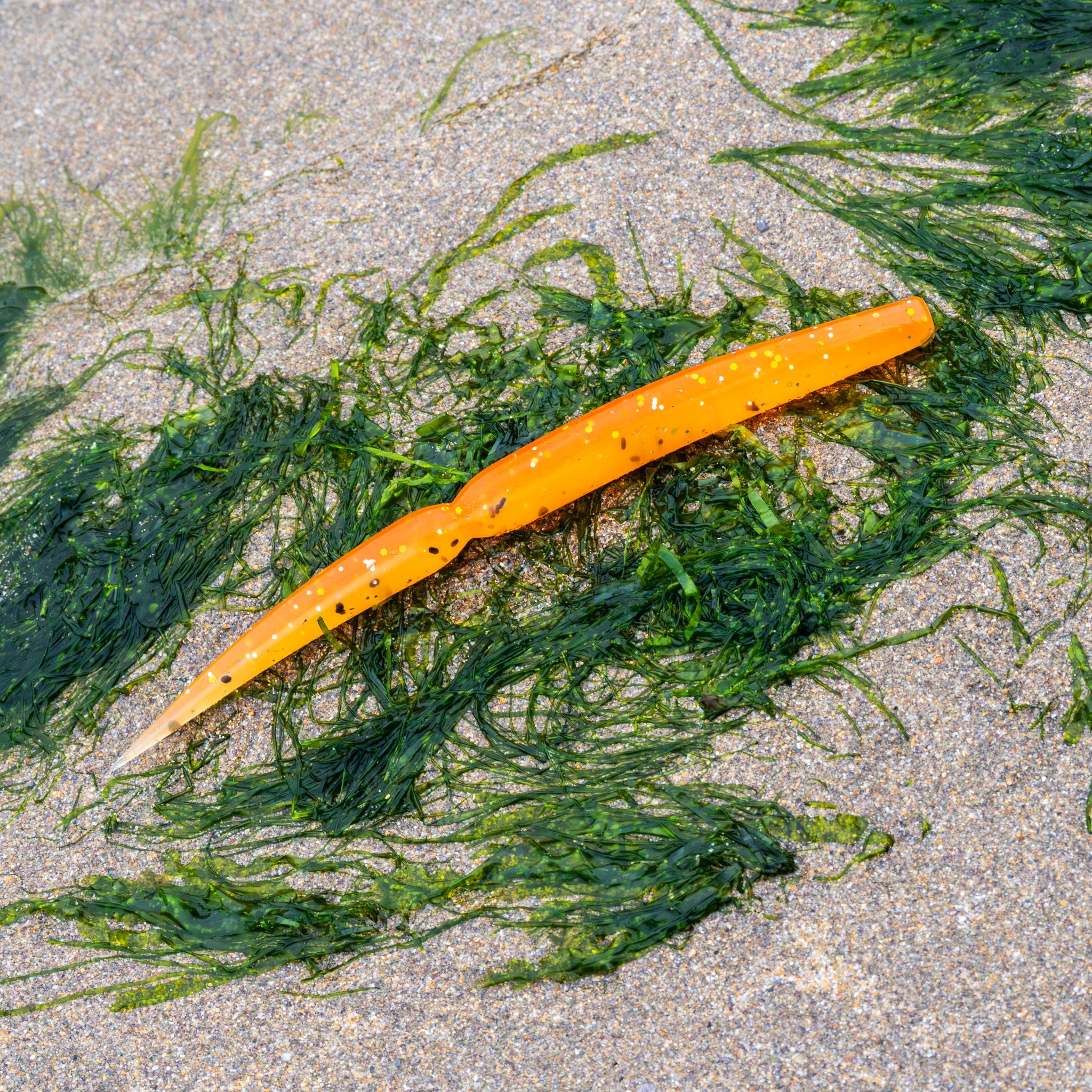 An orange soft plastic fishing lure with black glitter flecks, shown on a sandy beach surrounded by green seaweed.