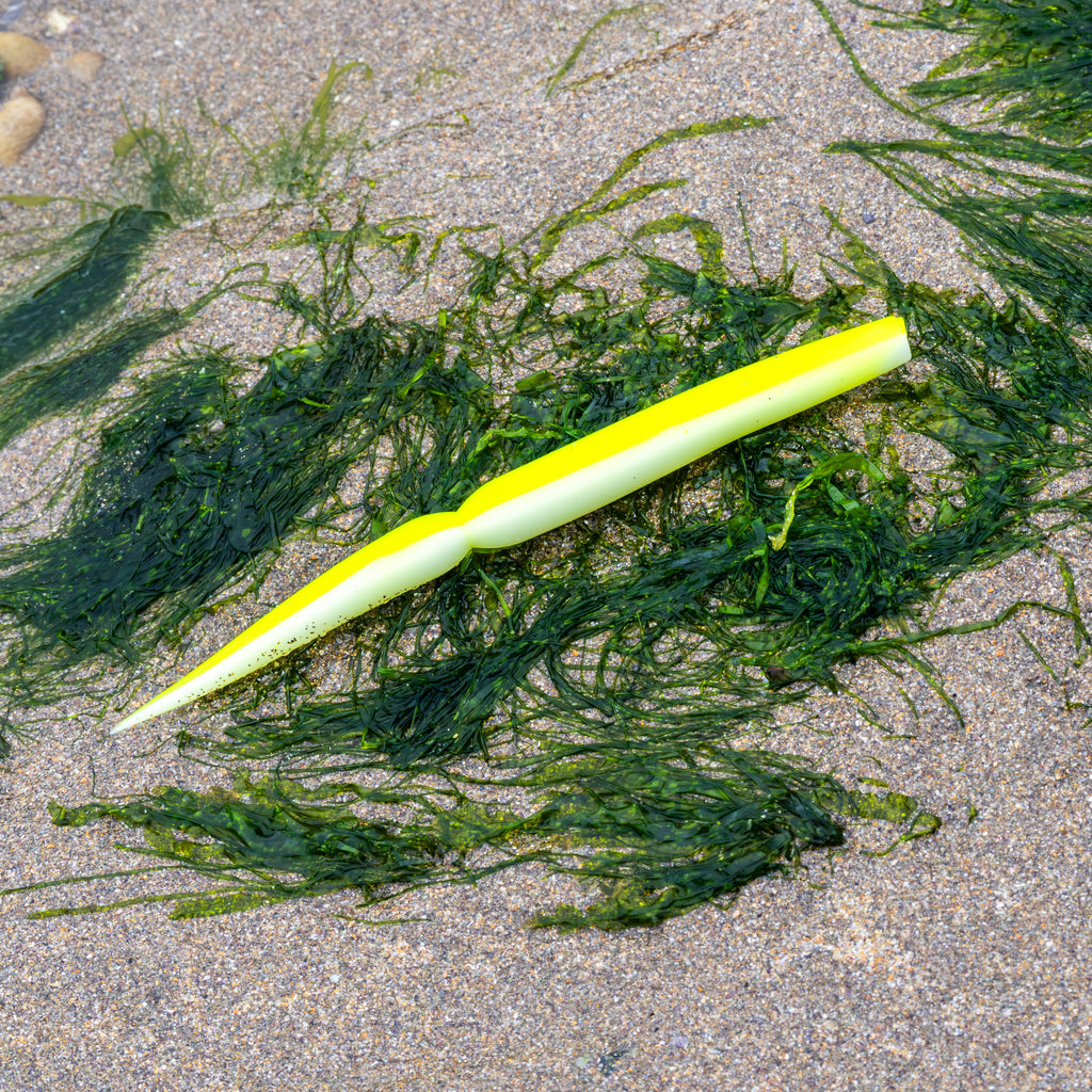A bright yellow and white silicone fishing lure with a ribbed body, lying on a sandy beach surrounded by green seaweed.