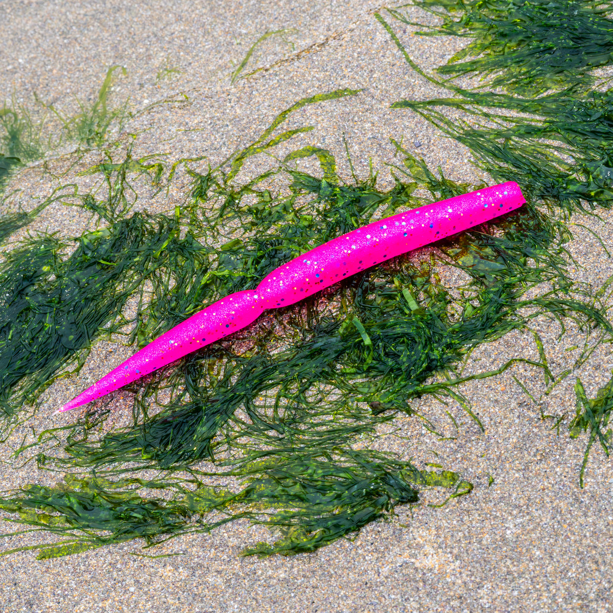 A bright pink soft plastic fishing lure with black glitter speckles, laid diagonally on a sandy beach surrounded by green seaweed.