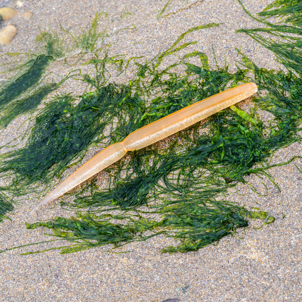 A silicone fishing lure with a ribbed body, lying on a sandy beach surrounded by green seaweed.